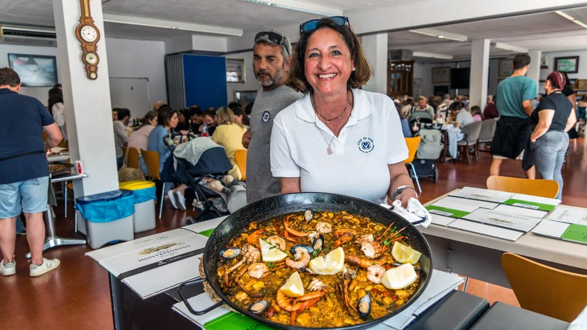 Mujer sonriendo con delantal, sosteniendo una paella con mariscos y limón en un restaurante concurrido.