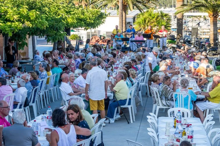 Concierto al aire libre con muchas personas mayores sentadas en mesas, música en vivo con músicos y cantantes en un escenario.