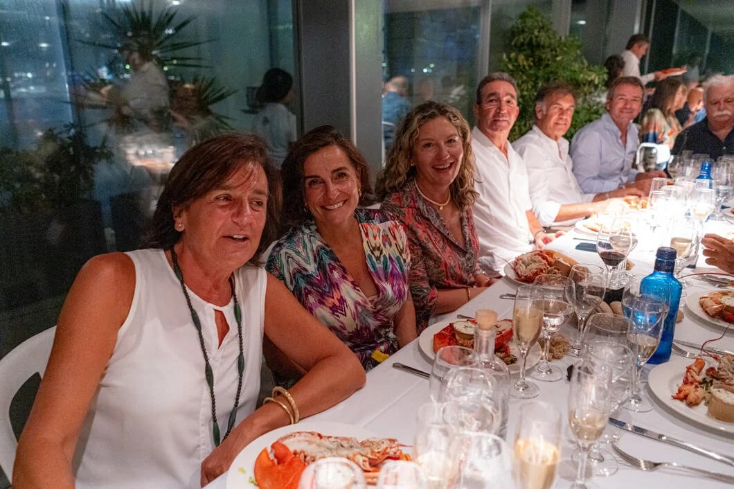 Grupo de seis personas con sonrisas en una cena en un restaurante, con alimentos y bebidas en la mesa, ambiente interior con ventanas y plantas decorativas.