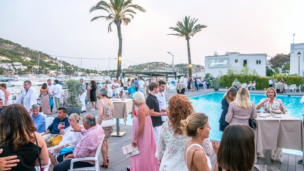 Reunión social en un evento junto a la piscina, con personas conversando y disfrutando, con vista a yates y montañas en el fondo.