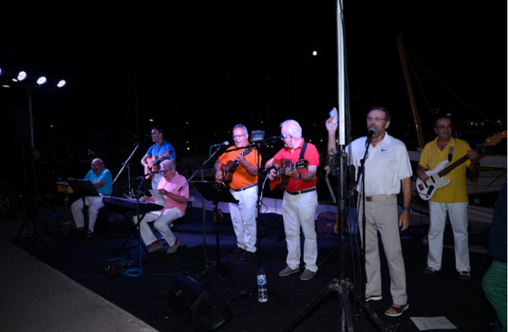 Grupo de músicos tocando en un escenario al aire libre en la noche, con instrumentos como guitarra, teclado y bass, frente a un muelle y yates.