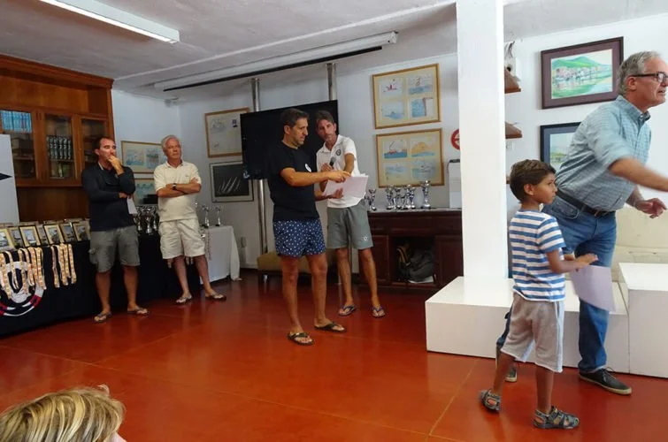 Un grupo de personas en una sala, aparentemente en una ceremonia o evento, con diplomas y premios en exhibiciones a la izquierda.