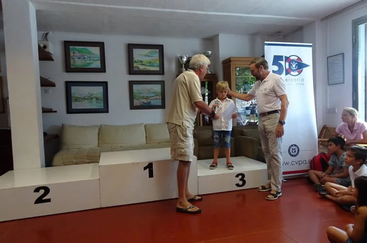 Niño en el podio recibiendo un premio de dos adultos en un evento de premiación, con otros niños sentados a un lado.