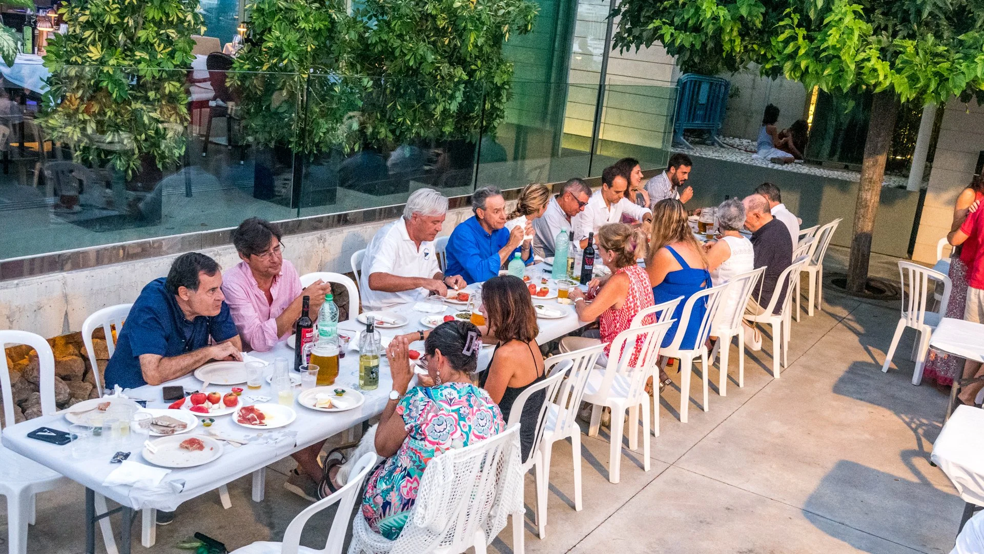 Grupo de personas cenando al aire libre en una reunión social en un restaurante, con comida y bebida en la mesa, rodeados de árboles y vegetación.