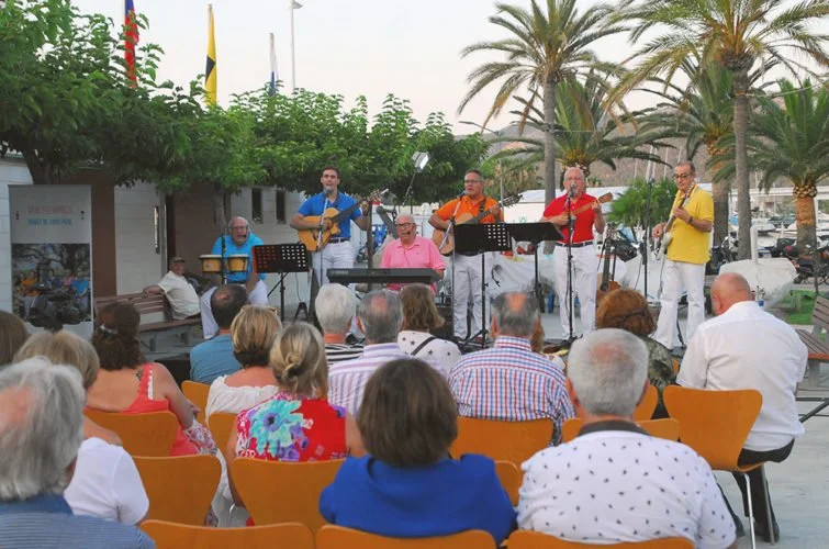 Un grupo de músicos tocando en vivo frente a una audiencia sentada, con palmeras y árboles en el fondo.