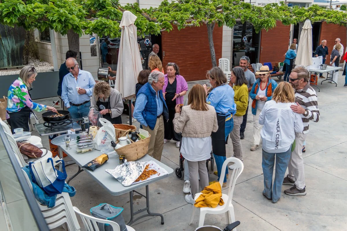 Grupo de personas en una reunión social al aire libre, con comida y bebidas en una mesa, niños y adultos charlando y disfrutando del evento.