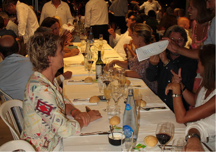 Personas sentadas en una mesa de banquet con comida y bebida, algunas riendo y otras conversando, en un evento social.