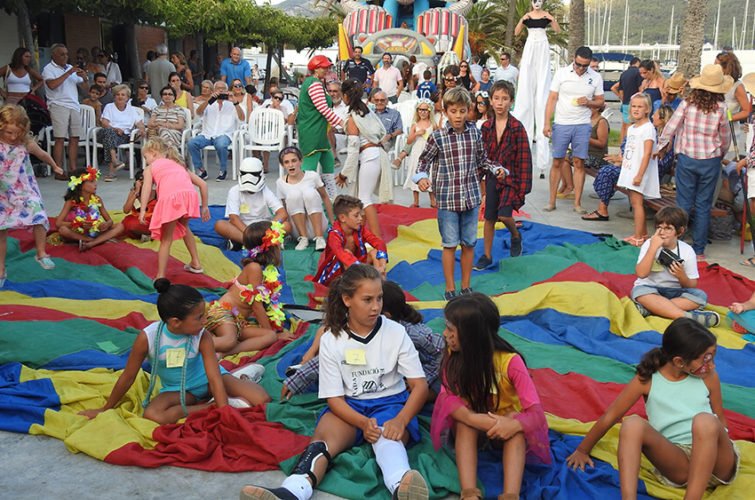 Niños y adultos en una celebración, con algunos niños disfrazados y en el centro un tapete grande de colores para juegos, en un ambiente festivo con inflables y personas sentadas observando.