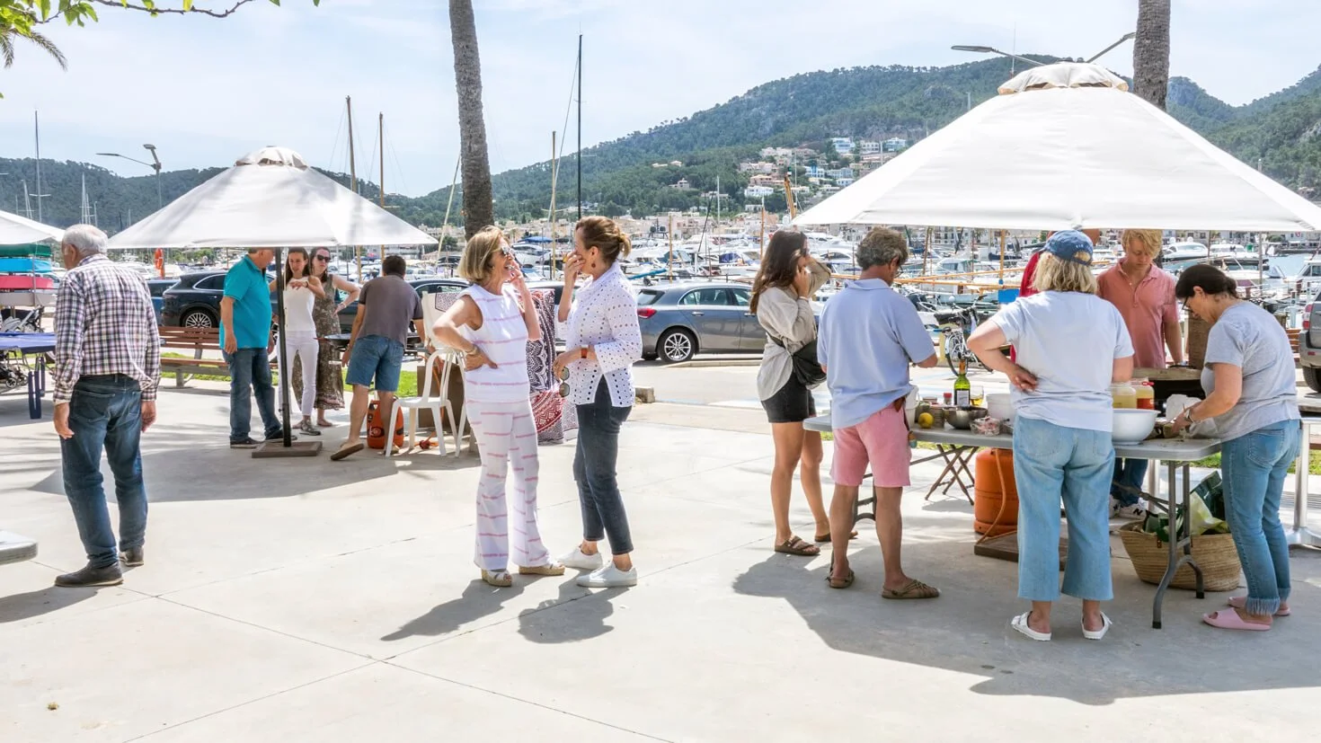 Personas en una reunión al aire libre cerca de un puerto con yates, comiendo y conversando bajo sombrillas, con montañas al fondo.