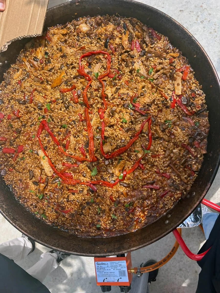Arroz frito con ingredientes como pimientos rojos, cebolla y otros vegetales en una sartén grande, encima de un quemador de gas portátil.