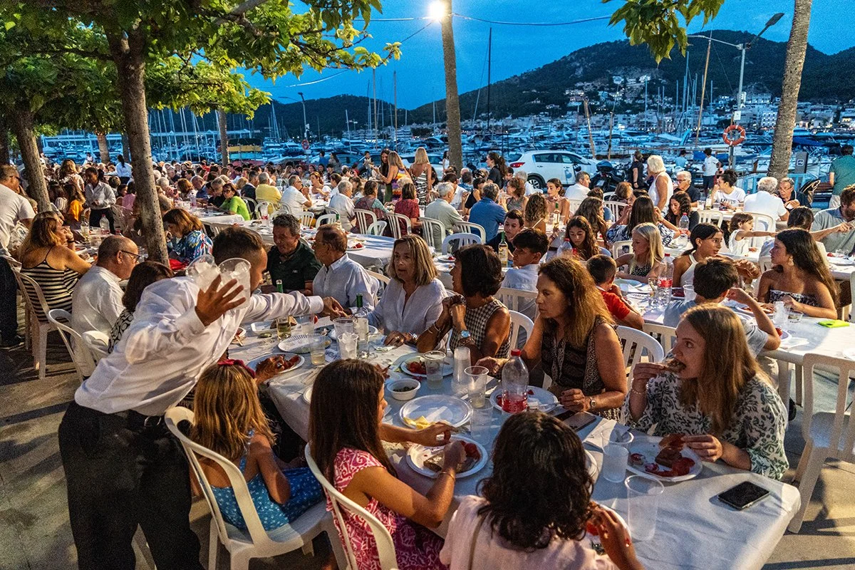 Cena al aire libre con muchas personas en una terraza junto a un puerto deportivo, disfrutando de una comida en la noche, rodeados de árboles y con montañas al fondo.