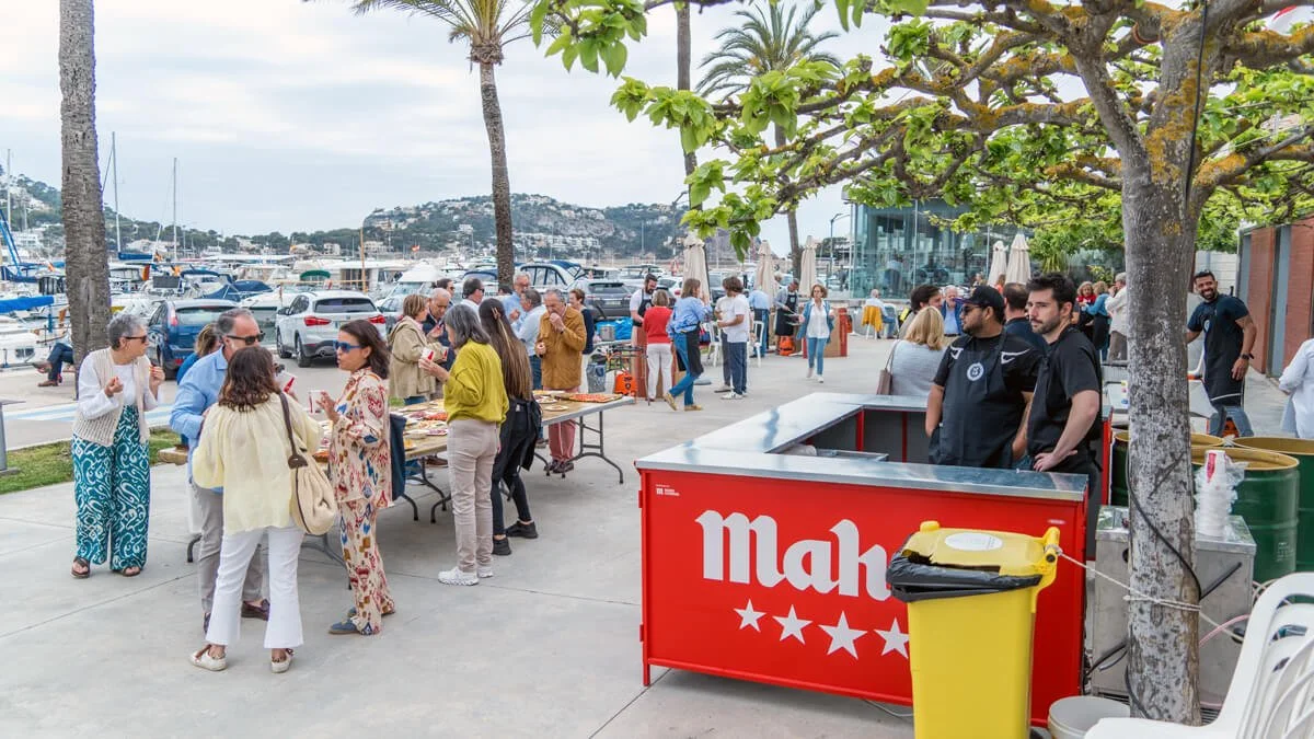 Personas en un evento al aire libre junto al puerto, conversando, sirviendo comida y disfrutando del día en un área con árboles y vistas al mar.