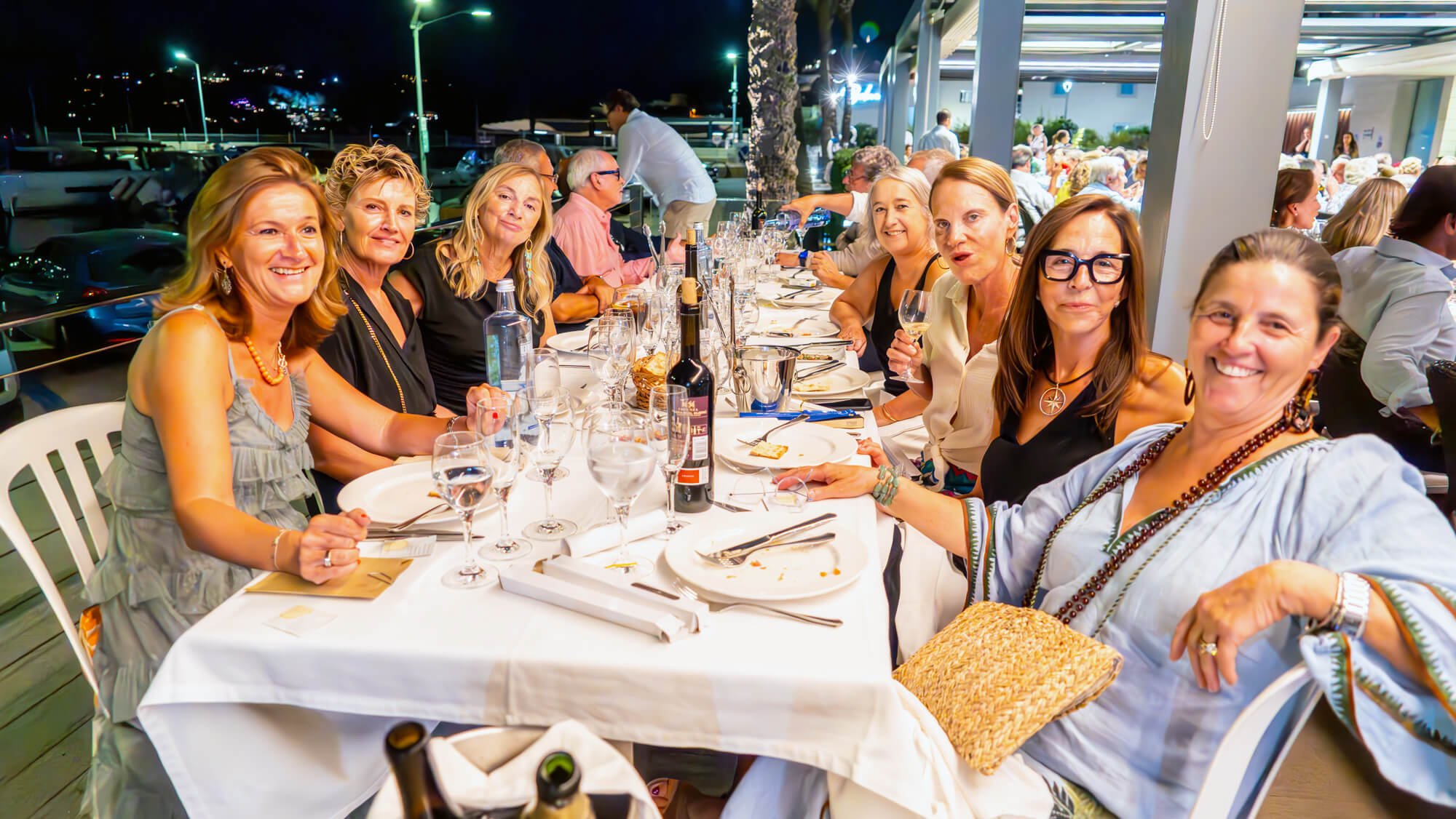 Grupo de personas en una cena al aire libre en la noche, sentados en una mesa con comida y vino, disfrutando de una celebración