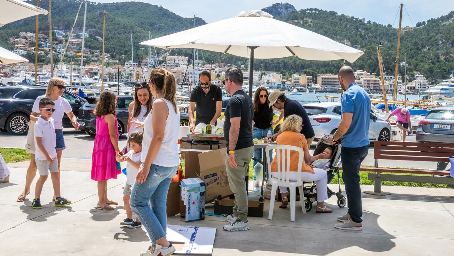 Grupo de personas en un muelle cerca del mar, participando en una comida o picnic, con yates y casas en las colinas de fondo.