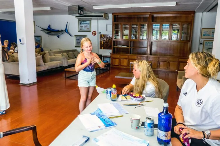 Una niña pasa un pastel a una mujer que está sentada en una mesa con varias personas, en un salón con decoracion marina y muebles de madera.