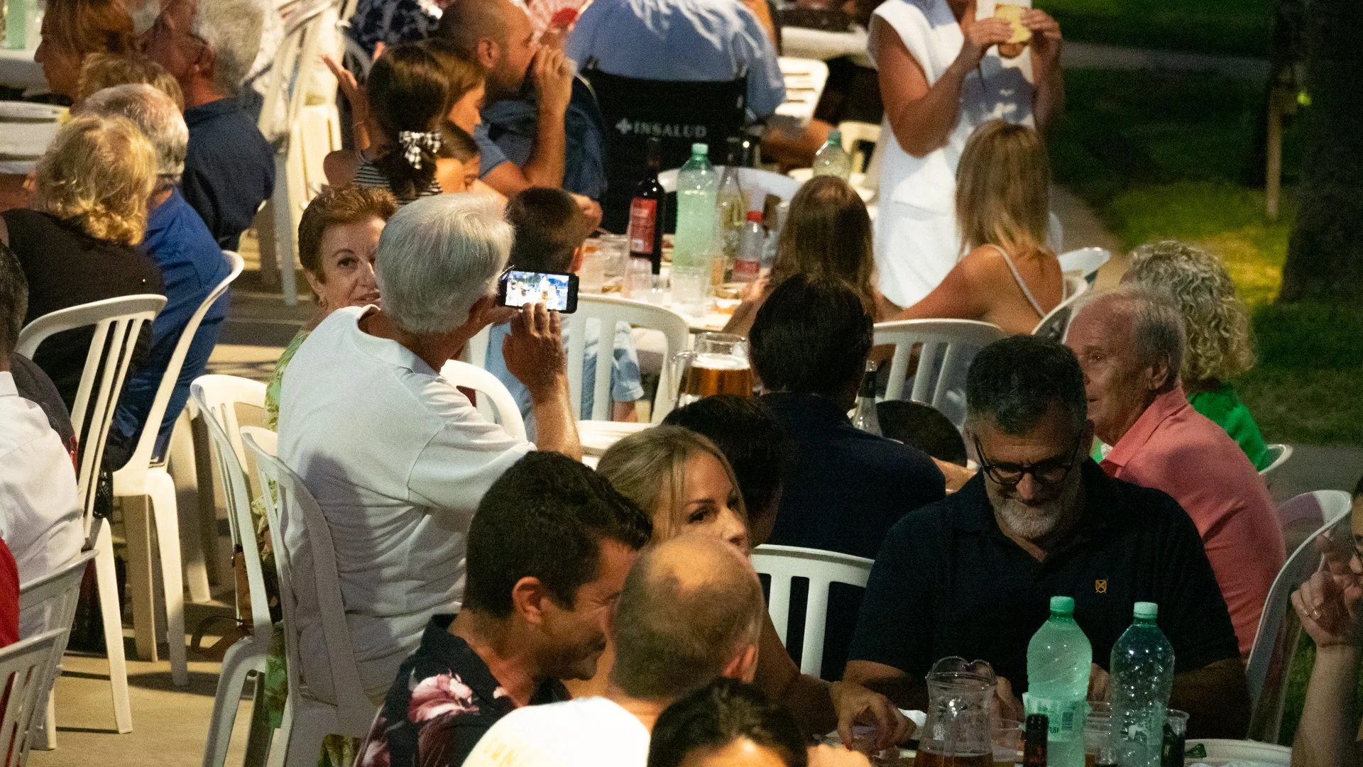 Grupo de personas sentadas en una mesa al aire libre, compartiendo una comida, algunas tomando fotos y otras conversando.
