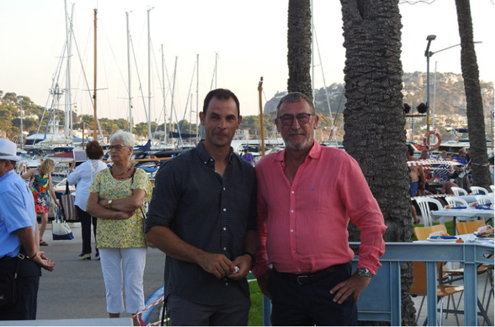 Dos hombres posando juntos en un muelle con yates, palmeras y gente en el fondo