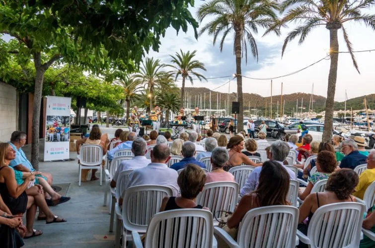 Personas asistiendo a un concierto al aire libre junto a un muelle con yates y palmeras, en un día soleado y despejado.