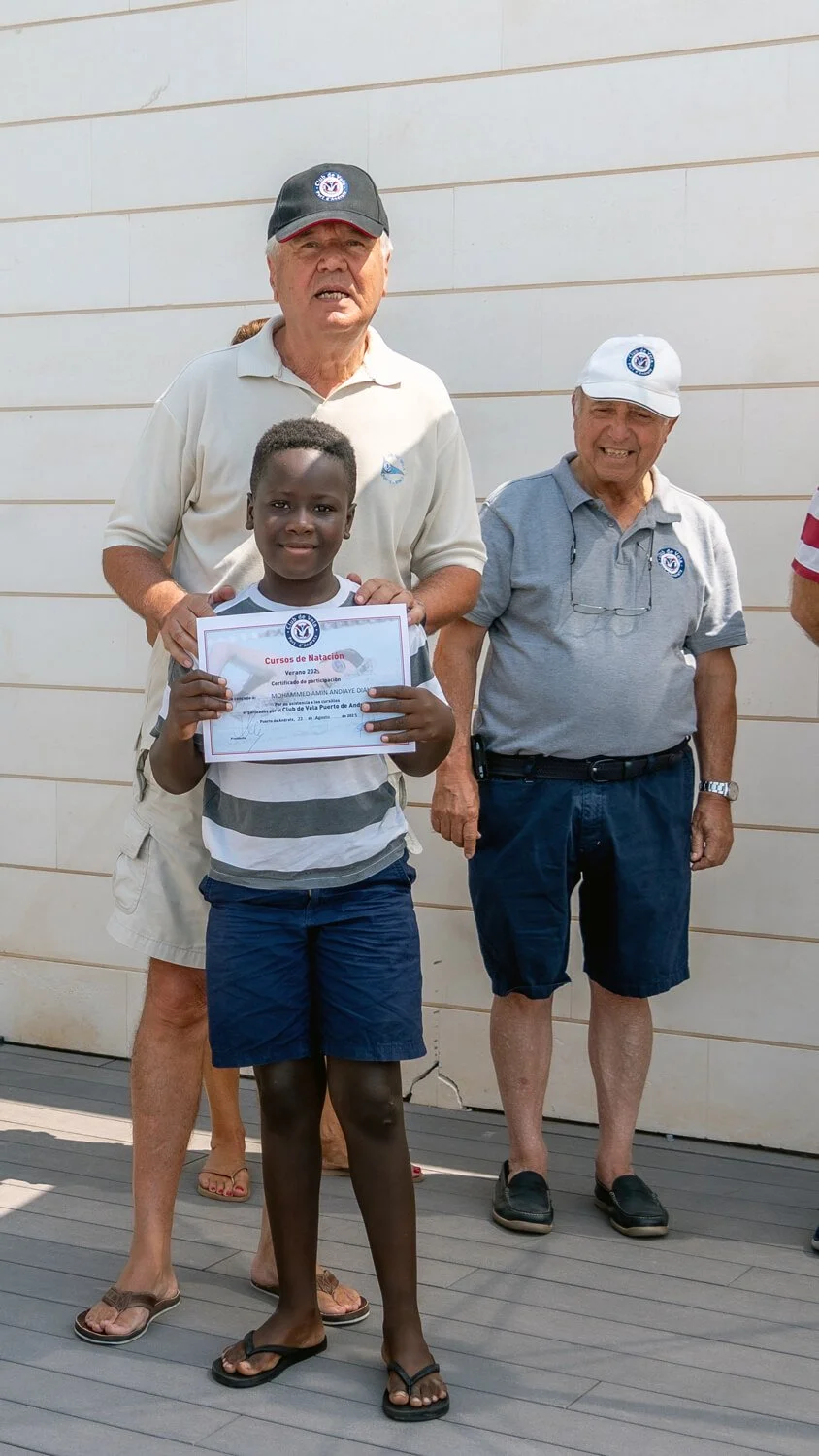Un niño sostiene un certificado de participación en un curso de natación, rodeado por tres adultos con camisetas y gorras, en un fondo de pared de color claro.