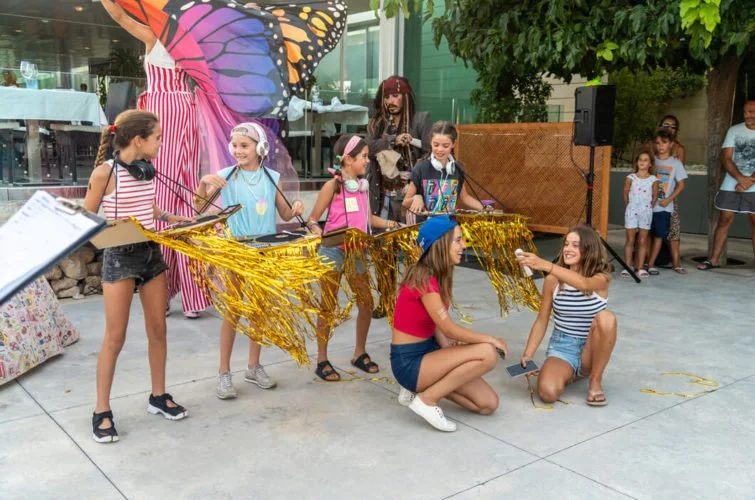Niñas en un evento al aire libre, con disfraces y decoraciones doradas, participando en actividades y música, mientras una persona en un disfraz de mariposa gigante observa.