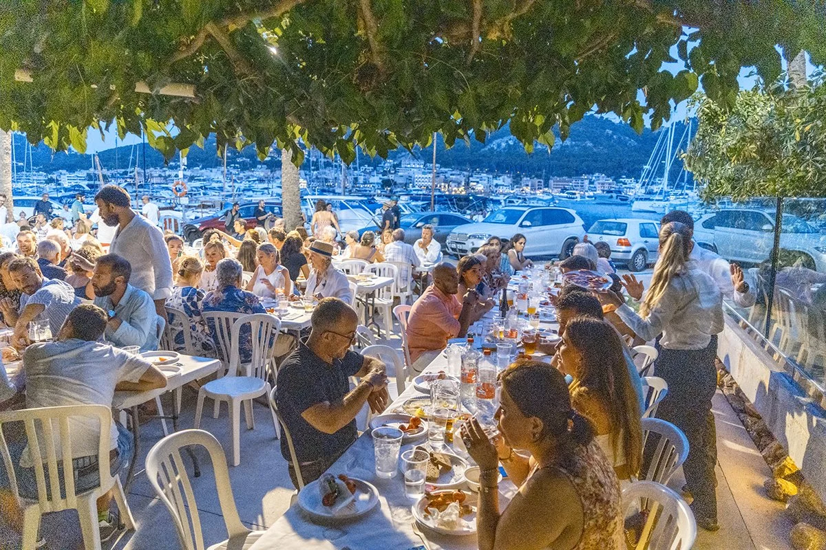 Una comida al aire libre en un restaurante junto a un muelle con muchas personas sentadas en mesas, comiendo y conversando, con barcos y un puerto en el fondo.
