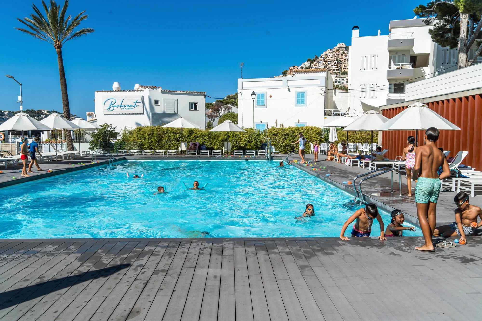 Personas disfrutando en una piscina al aire libre con sombrillas blancas y edificios blancos de fondo, en un día soleado con cielo azul.