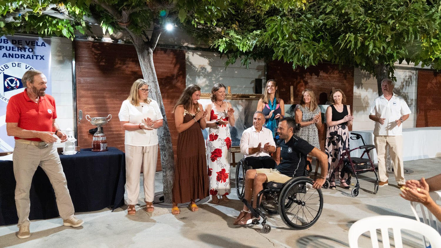 Personas en ceremonia de premiación, con una persona en silla de ruedas y otra con caminador, en un espacio al aire libre con árboles, mesas y un banner de club de vela.