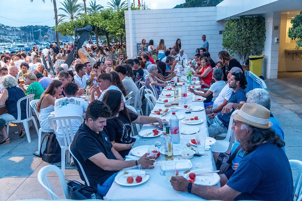 Reúna de personas en una fiesta al aire libre, sentados en una larga mesa con comida y bebidas, con un fondo de palmeras, y yates en el puerto.