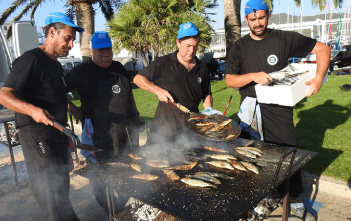 Cuatro hombres cocinando pescado a la parrilla en un parque, con palmas y barcos en el fondo, todos con camisetas negras y gorros azules.