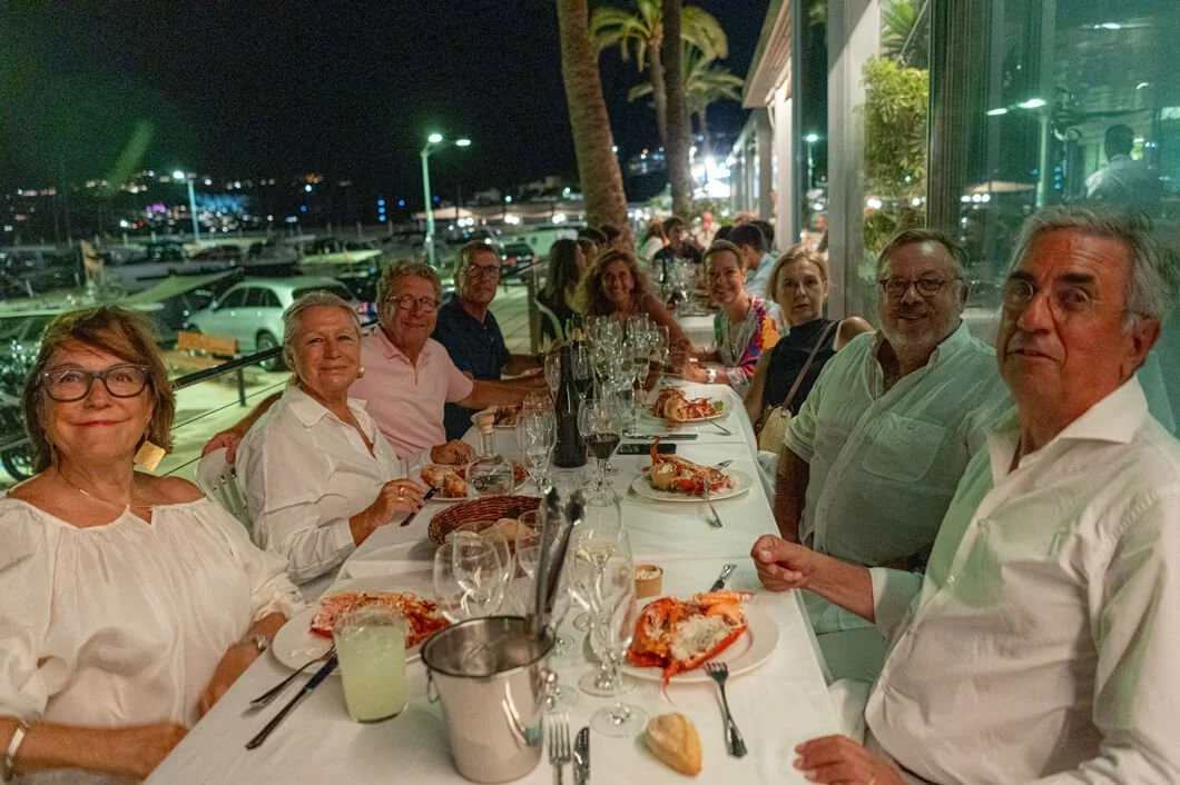 Grupo de personas cenando en un restaurante junto a un puerto con yates, en la noche.