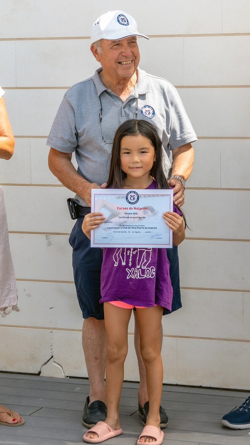 Niña sosteniendo certificado en un evento de natación, acompañada por un adulto, en un escenario al aire libre.