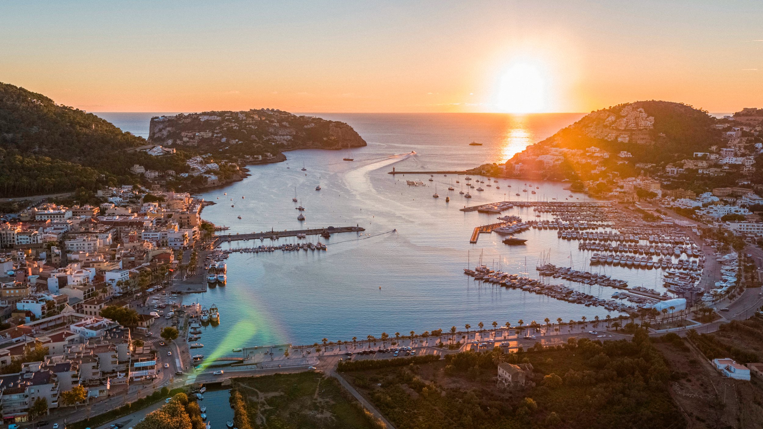 Vista aérea de un puerto en una ciudad costera al atardecer, con barcos atracados y montañas en los alrededores.