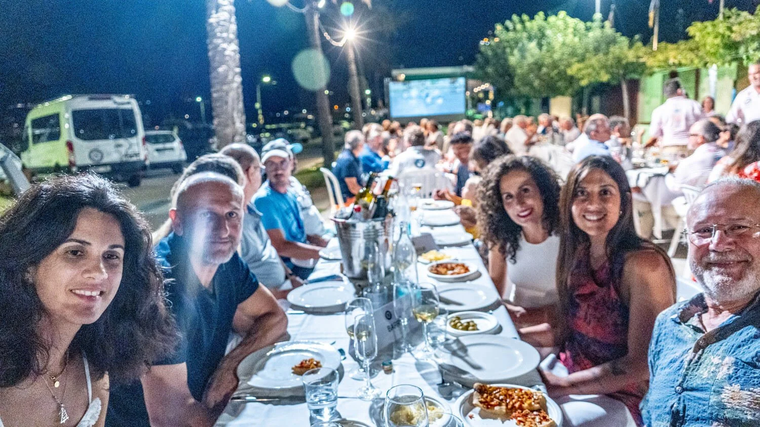 Grupo de personas cenando en una mesa al aire libre en la noche, con comida y bebidas en una fiesta o reunión social.
