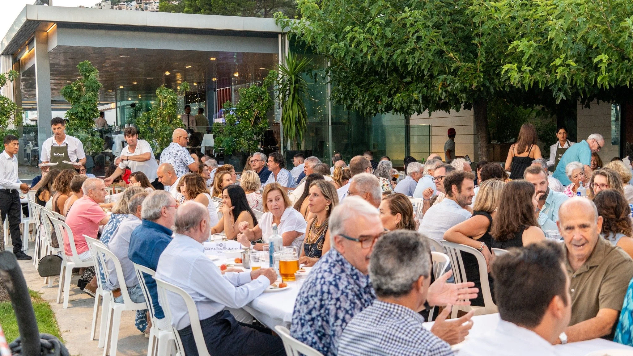 Una multitud de personas sentadas en una mesa al aire libre durante un evento social, con vegetación y un edificio moderno en el fondo.