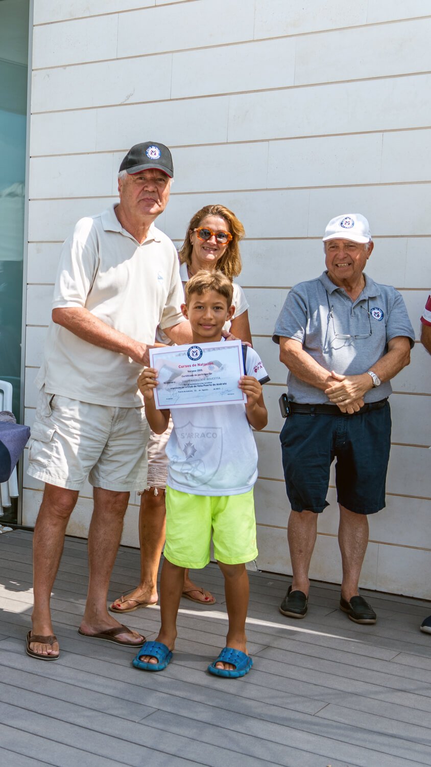 Niño sosteniendo un certificado de clases de natación, rodeado de tres adultos, en un evento al aire libre, con paredes blancas de fondo.