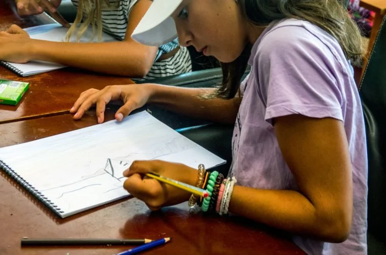 Niña en una mesa con cuaderno y lápiz dibujando, con otras personas alrededor.