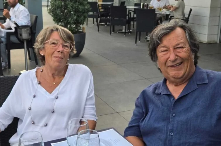 Dos personas mayores, una mujer con gafas y una blusa blanca, y un hombre con camisa azul, sentados en una mesa de restaurante al aire libre, sonriendo.