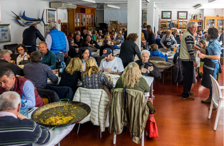Personas sentadas en mesas en un restaurante, sirviéndose comida, conversando, y disfrutando del ambiente interior.