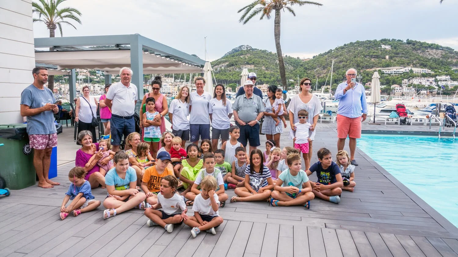 Grupo de niños y adultos posando frente a una piscina con fondo de mar y colinas verdes en un ambiente de playa