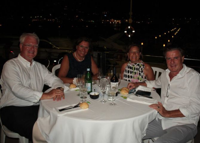 Cuatro personas sentadas en una mesa de comida en un restaurante o barco, noite, con vista al agua y luces de fondo.