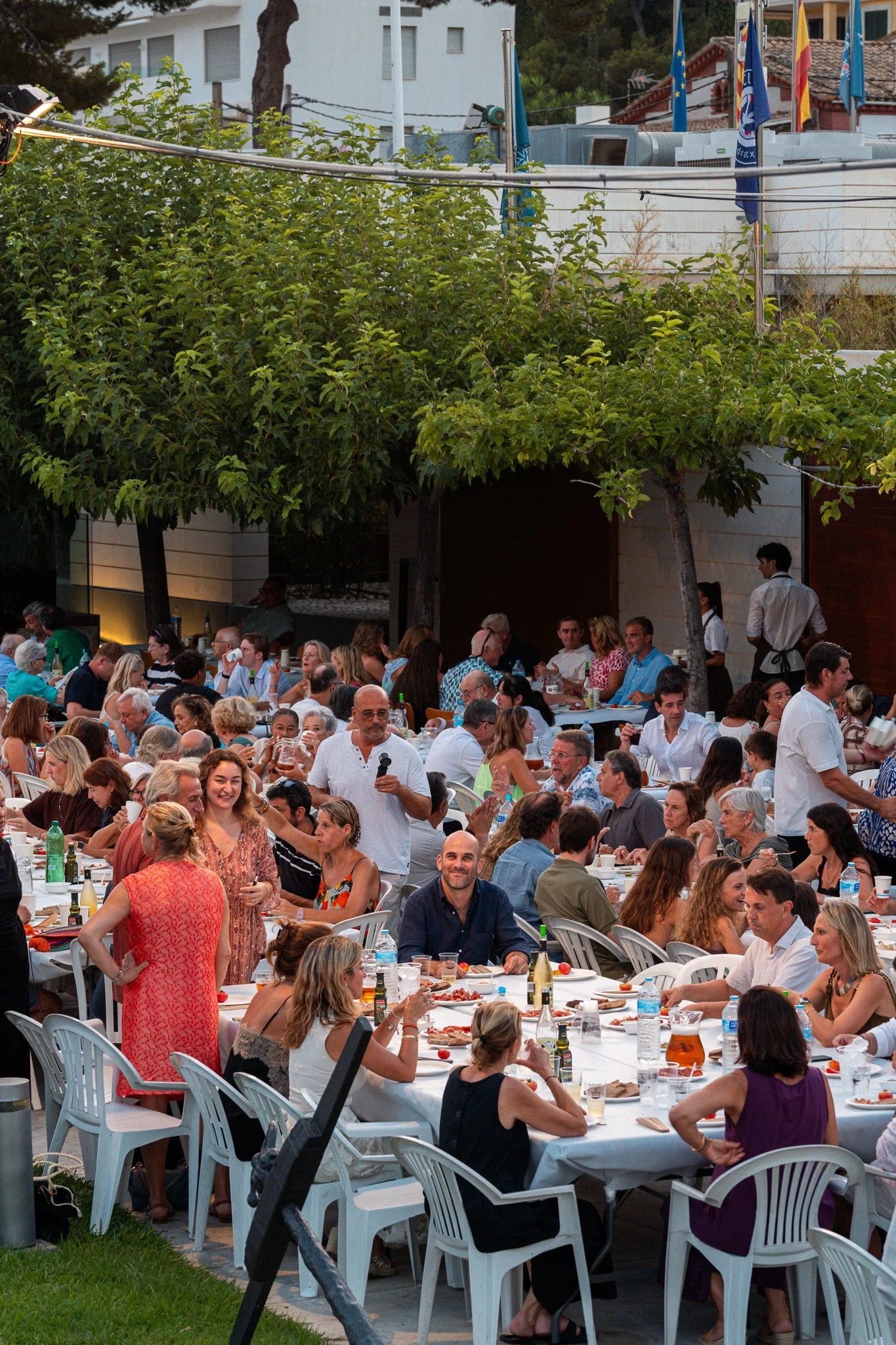 Gran reunión al aire libre con muchas personas comiendo y socializando, mesas cubiertas con manteles blancos y decoración festiva, árboles y banderas en el fondo, ambiente de celebración vespertina.