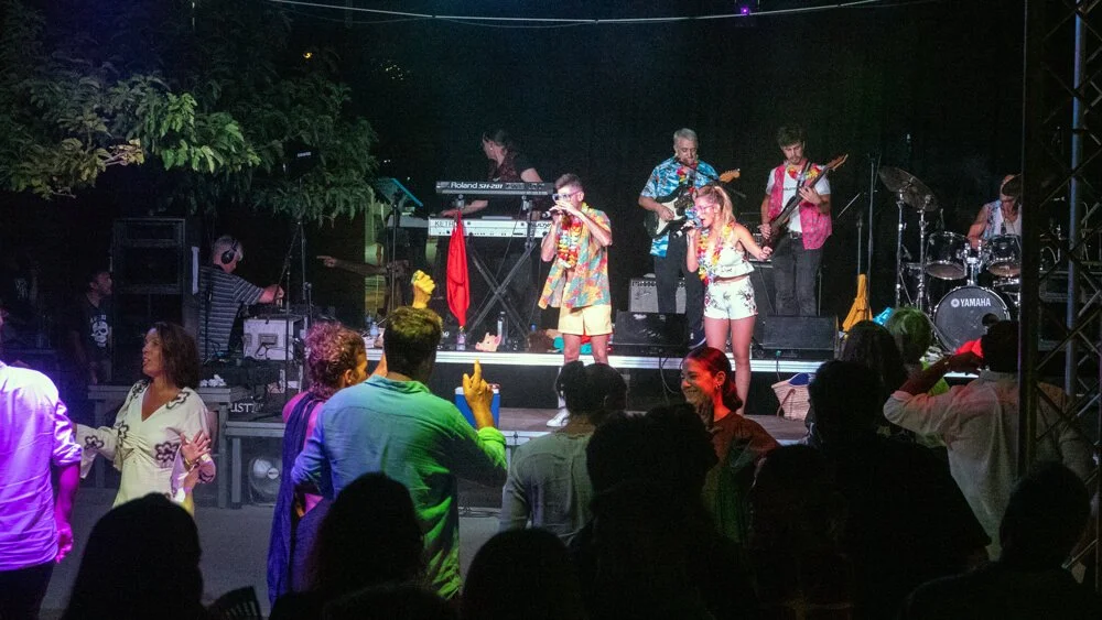 Un grupo de músicos en un escenario tocando instrumentos y una multitud disfrutando de un concierto al aire libre en la noche, con luces y árboles en el fondo.
