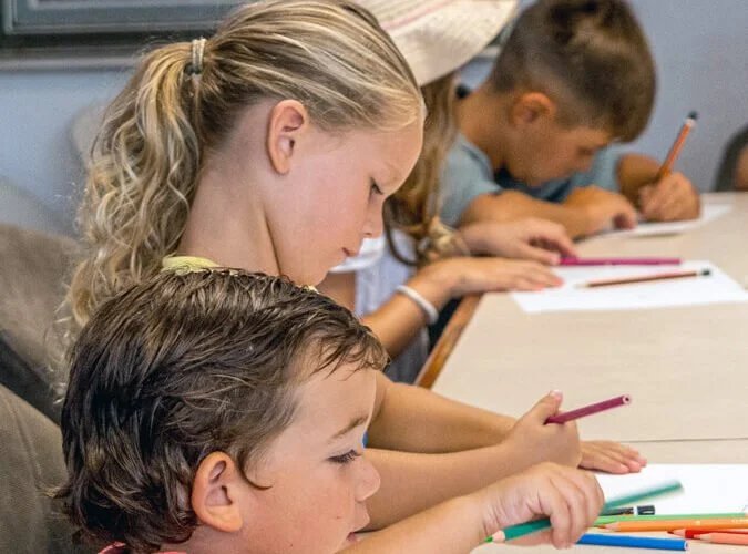 Niños sentados en una mesa escribiendo en hojas de papel con lápices de colores.