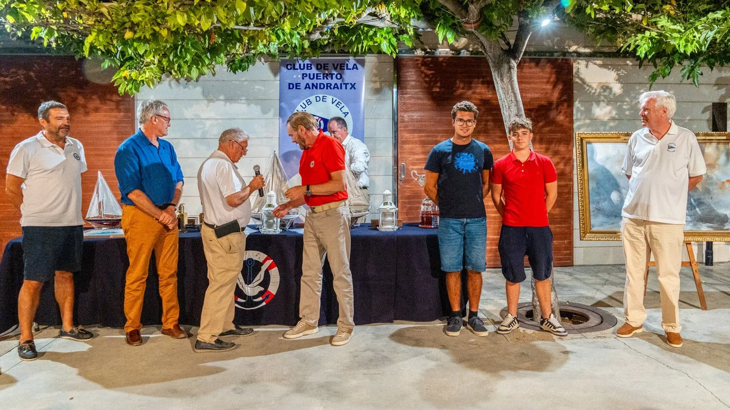 Grupo de personas recibiendo un reconocimiento en una ceremonia junto a un cartel de club de vela. Al fondo, un árbol y dos cuadros en exhibición.