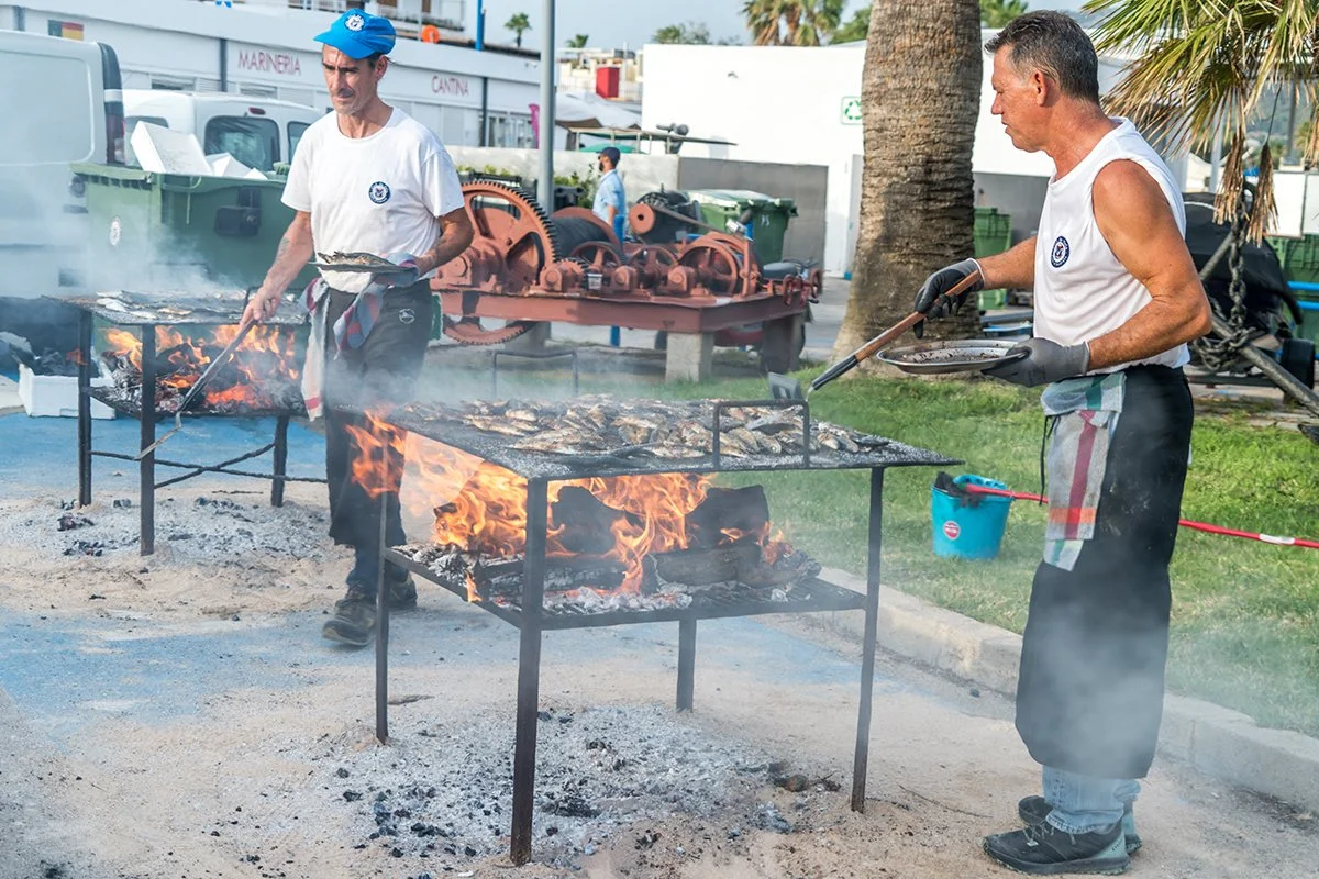 Dos hombres cocinando en una parrilla con fuego abierto en la calle, mientras usan guantes y tratan la carne.