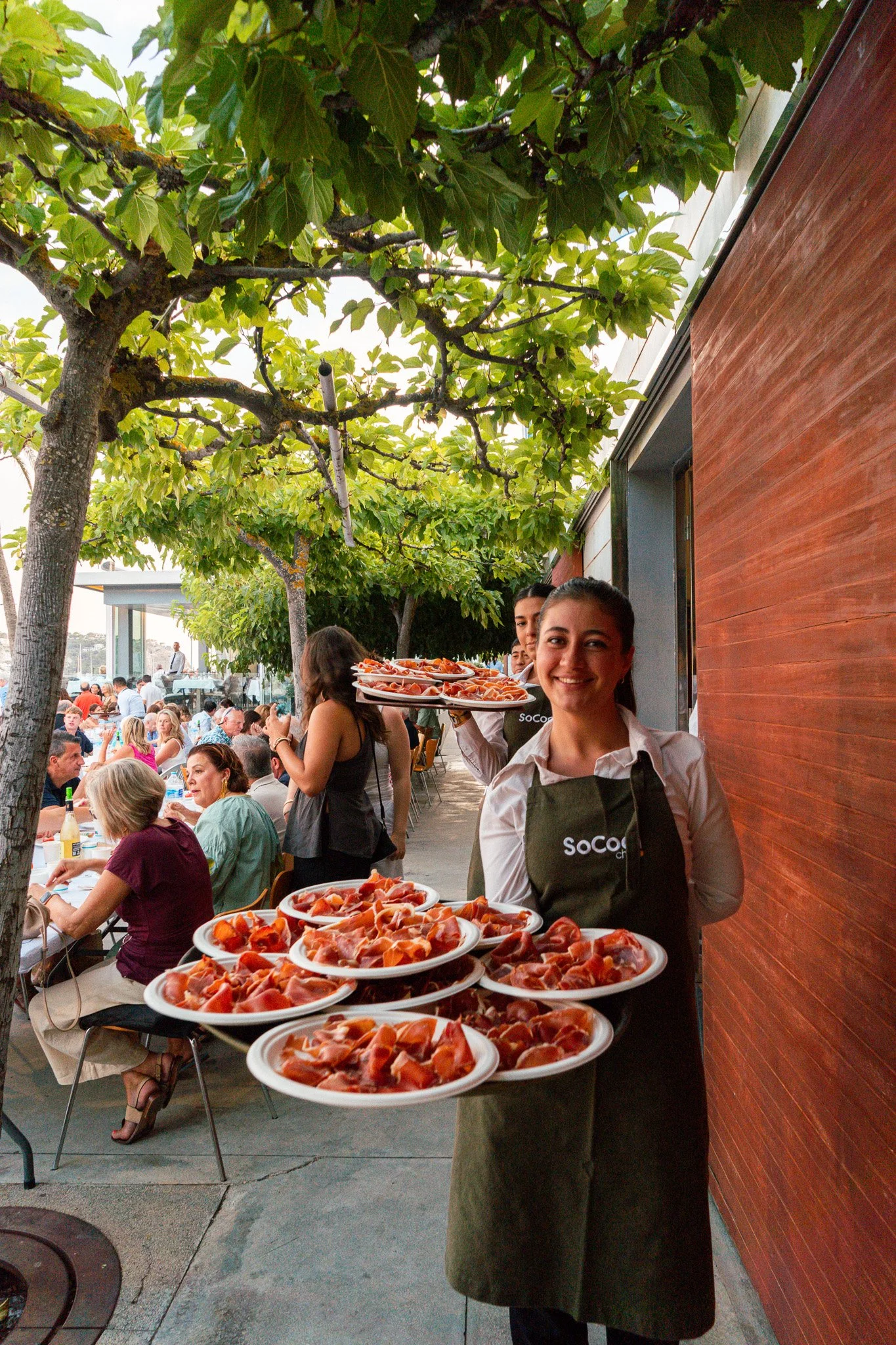 Mesera sonriendo con delantales verdes que dicen 'SoCo', presentando platos de jamón en una comida al aire libre con muchas personas sentadas en mesas.