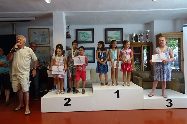 Niños en un podio de premiación, con medallas y certificados, en un evento de reconocimiento, con adultos y cuadros en la pared en el fondo.