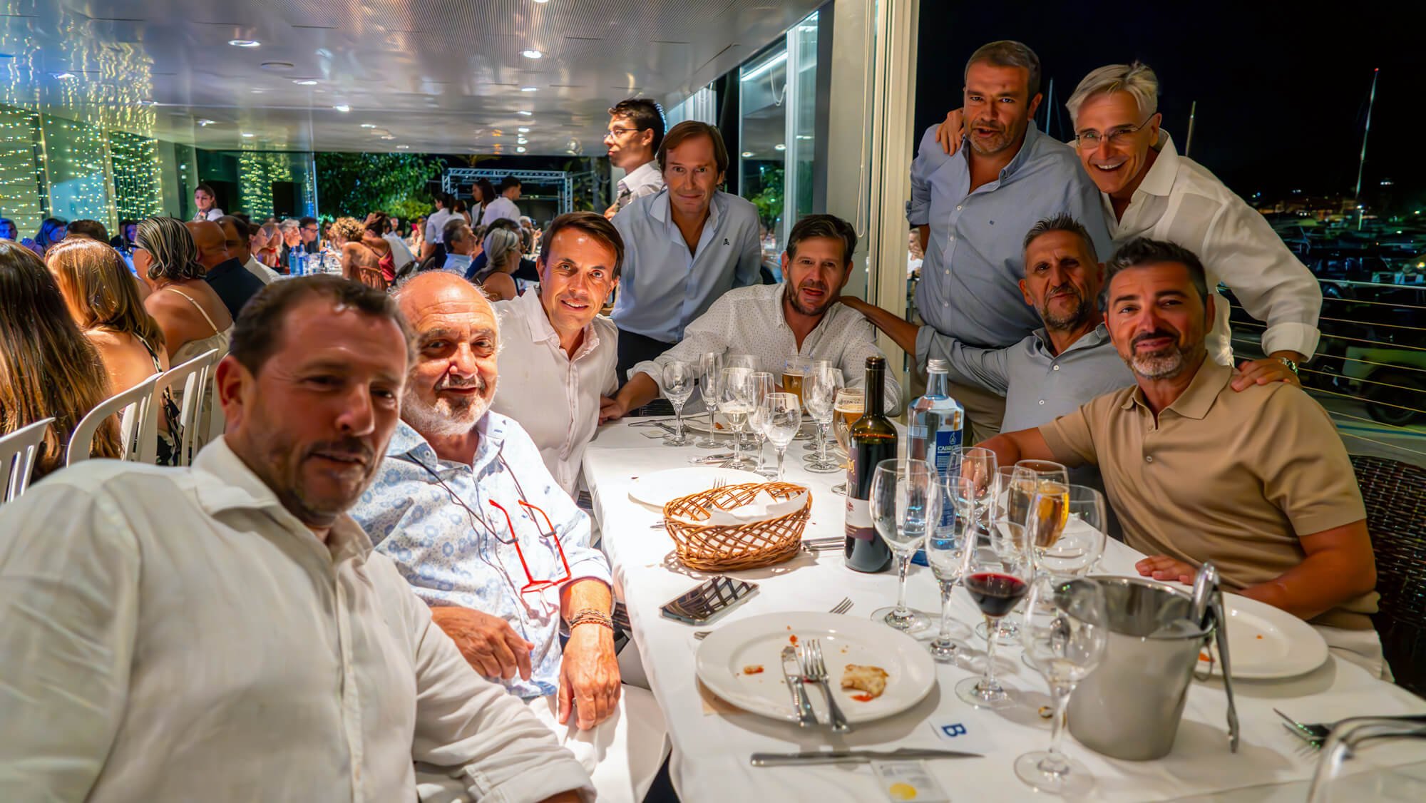 Grupo de personas sentadas en una mesa de restaurante durante una cena, con varias bebidas en copa y botella, en un ambiente iluminado y con vista a la noche.