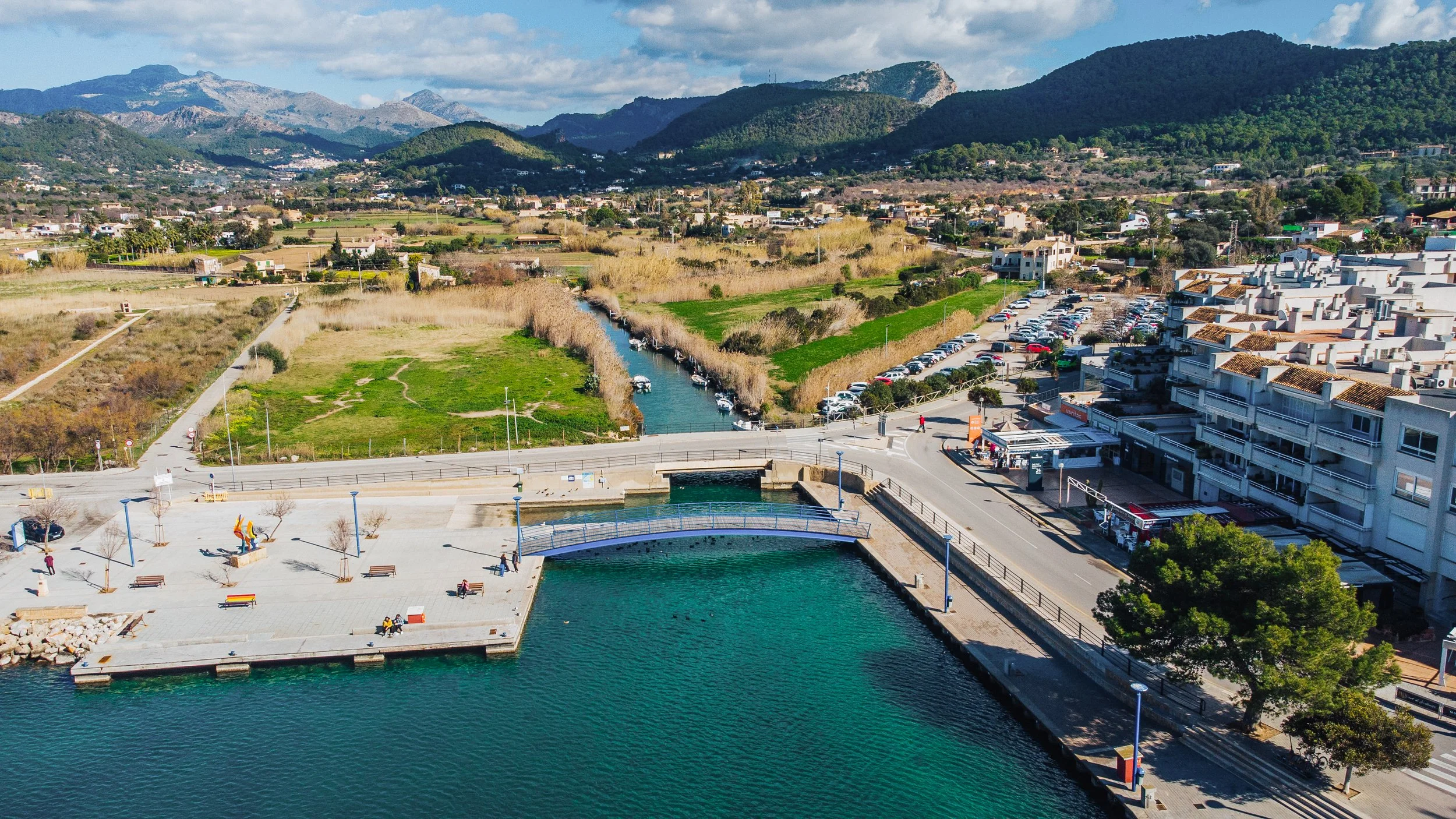 Vista aérea de un río que atraviesa una zona urbana con edificios, calles y una plaza con bancos y esculturas, rodeada de áreas verdes y montañas al fondo.