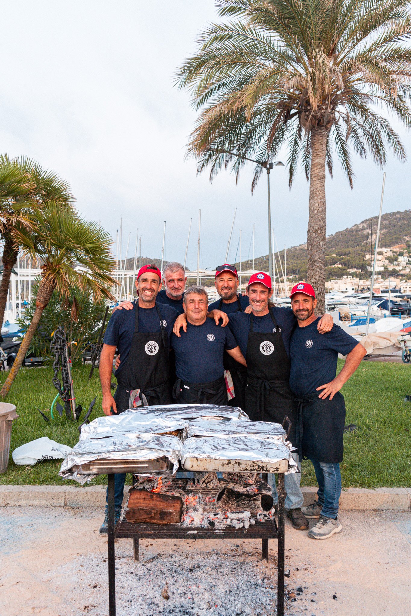 Grupo de siete hombres sonriendo en un evento al aire libre junto a un asador con comida envuelta en papel aluminio. Hay palmeras, embarcaderos y colinas en el fondo.